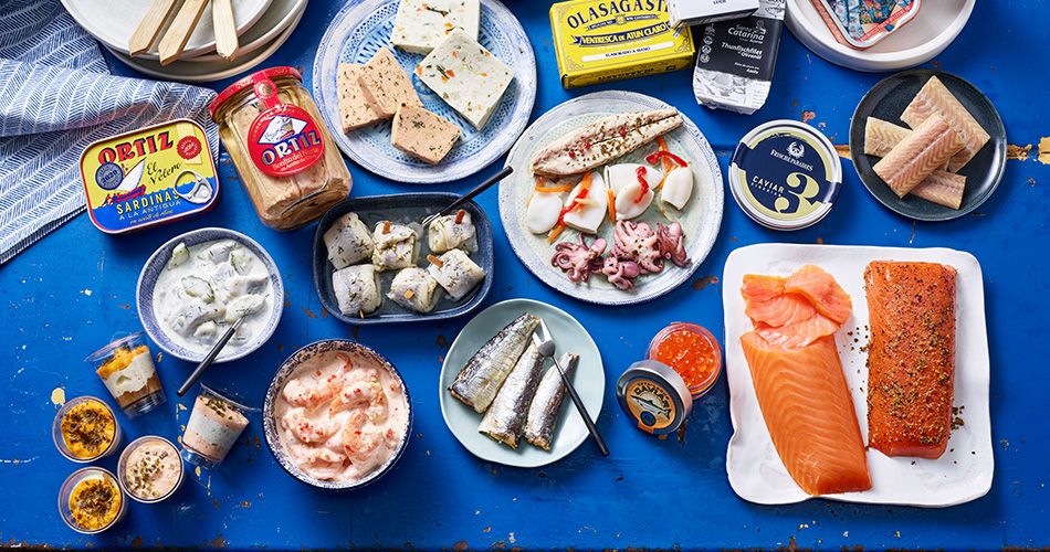 Kategorie Fischfeinkost Fischfeinkost in verschiedenen Ausprägungen wie Dosenfisch, Räucherfisch und Pasteten auf einem Tisch mit blauem Laken